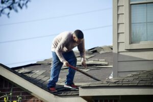 A worker on a residential roof uses a tool to remove old asphalt shingles.