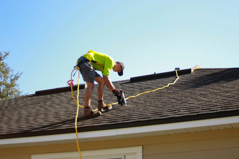 A male roofer for a roofing company, wearing a bright green shirt and shorts, is on a residential roof. He's working with a black and red nail gun, secured by a yellow safety harness and rope. The roof is covered with brown shingles under a clear blue sky.
